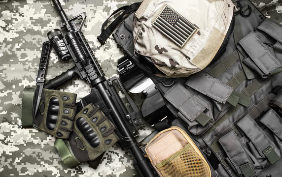 Upper view photo of a military tactacal bulletproof vest, gloves, helmet with american flag badge and rifle laying on camouflage cloth background.