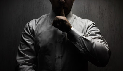 Photo of a scary horror man in white shirt standing under upper light on black background showing silence hand sign.