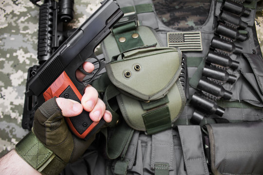 First person view hand in tactical gloves holding a gun on camouflage background with bulletproof vest and cartridge belt.