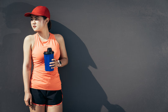 An asian woman athletic is jogging on the concrete road, she is warming her body and tideten her tying her shoes tightly fitting before workout. She is drinking the protain shak or cool water.