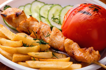 appetizing chicken kebab, fried potatoes chips,  grilled tomato and fresh sliced cucumbers close-up on white plate 