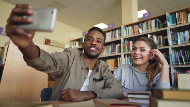 African merican guy and caucasian girl have fun smiling and taking selfie photos on smartphone camera at university library . Cheerful students have rest while prepare for examination