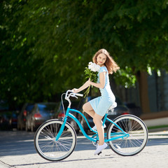 Naklejka na ściany i meble A young beauty on a vintage bicycle pinned her peonies on a blurry background of saturated urban greenery on a sunny day