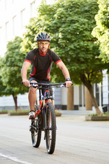 Obraz premium Man bicyclist in cycling garment and protective helmet, looking at camera, riding bike down empty city center alley surrounded by green trees. Sportsman training outdoors improving skills.
