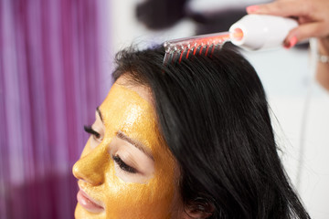 Closeup of hair treatment procedure performed to a brunette client of a beauty salon who has a gold mask on her face.