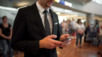 Businessman standing in airport hall, typing a message on phone, business trip