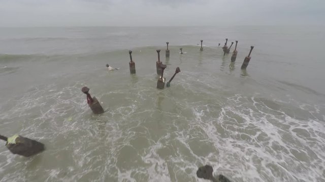 An aerial view of rusted iron pillars buried in the sand at the shore with waves crashing into them.