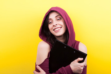 Smiling woman wearing a hood holds a black folder in hands on yellow background in studio