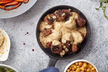 Flatlay with traditional swedish beef meatballs with gravy served in one portion black cast iron pan, handle wrapped in blue towel, plates with sauce, baked carrots and chickpeas. Dinner concept.