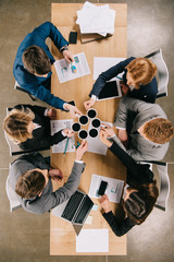 Top view of businesspeople with coffee cups in hands sitting at table with digital devices and documents