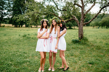 Fototapeta premium Three young girl Slavonic appearance in etno folk white traditional dresses outdoor summer portrait. Female friends with wreath of flowers on head celebrating feast at nature. Pagan style lovely women