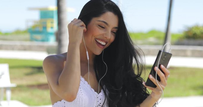 Pretty brunette using headphones and waving at camera of smartphone having videocall while chilling in summer park. 