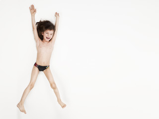 Smiling little boy in swimming trunks jumping with arms raised against a white background