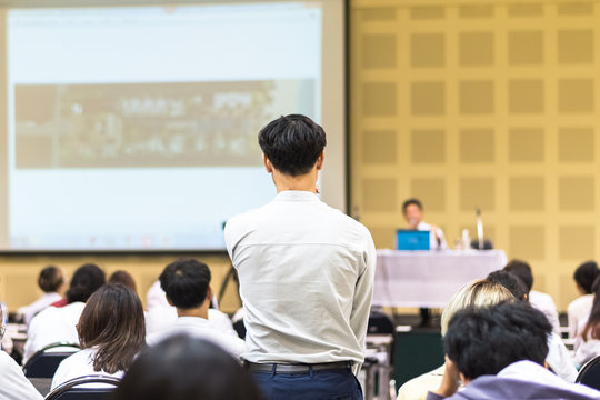 Seminar Q&A session with a participant rear view standing among audience group participating in  asking question to speaker for answer in entrepreneur business conference or education lecture hall