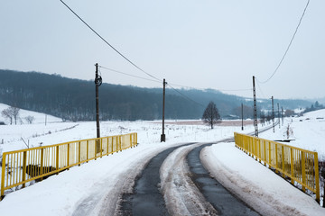 Obraz premium snowy winter landscape, asphalt road across yellow bridge leads in to the hills, pylons and wires follow the path