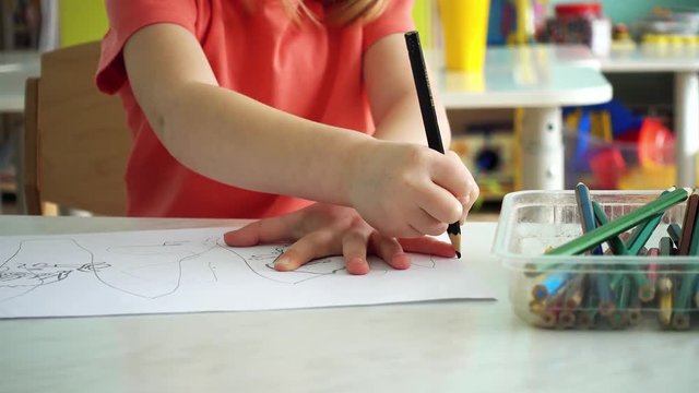 A child in a kindergarten. The girl draws pencils for 4-5 years. In the background are shelves with toys.