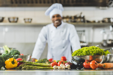 Fototapeta premium smiling african american chef at restaurant kitchen with vegetables on foreground
