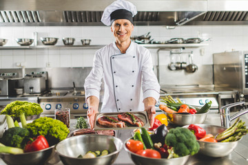 smiling chef taking tray with raw meat at restaurant kitchen