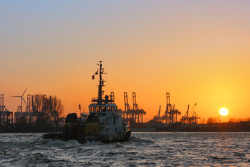 Tugboat at sunset on the Elbe river. Silhouettes of a shipyard cranes in the port of Hamburg