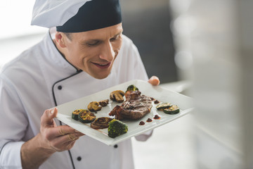 Obraz na płótnie Canvas handsome chef sniffing cooked steak with vegetables at restaurant kitchen