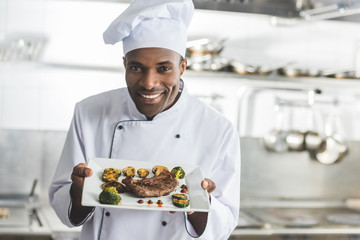happy african american chef holding plate with cooked steak and vegetables at restaurant kitchen