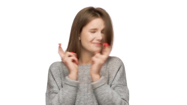 Portrait of concentrated woman dressed in basic sweatshirt willing good luck and pleading with keeping fingers crossed over white background in studio. Concept of emotions