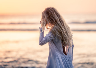 Beautiful blonde girl with long hair in short white dress walking at sunset on the beach in Bali,...