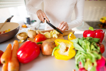 Young woman is cutting tomatoes and potatoes and preparing breakfast while standing in front of a kitchen counter filled with fresh vegetables.
