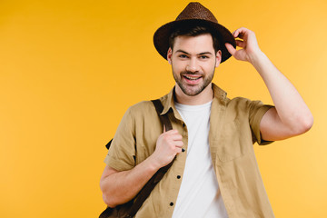 handsome young man adjusting hat and smiling at camera isolated on yellow