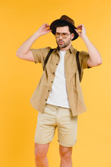 portrait of handsome young man in sunglasses and shorts adjusting hat isolated on yellow