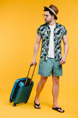 handsome young man in summer outfit holding suitcase and looking away isolated on yellow