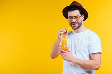 Fototapeta premium handsome young man in hat and sunglasses drinking summer cocktail and smiling at camera isolated on yellow
