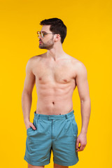 handsome shirtless young man in shorts and sunglasses standing with hand in pocket and looking away isolated on yellow