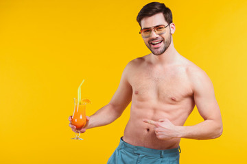 handsome shirtless young man in shorts holding glass of summer cocktail and pointing with finger...