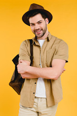 handsome young man in hat standing with crossed arms and smiling at camera isolated on yellow