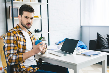 Young man holding coffee cup by table with laptop at home office