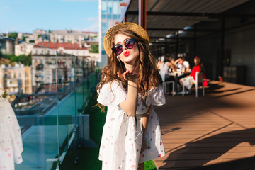 Pretty girl  in hat with long hair is listening to music through headphones on the terrace. She wears a white dress with bare shoulders, sunglasses, red lipstick. She is sending a kiss to camera.