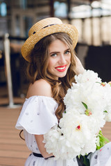Vertical close-up portrait of  girl with long hair in hat sitting on floor on the terrace. She wears a white dress with naked shoulders, red lipstick. She holds has white flowers in hands.
