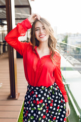 Pretty girl with long hair is posing on terrace. She wears red shirt and black skirt. She is...