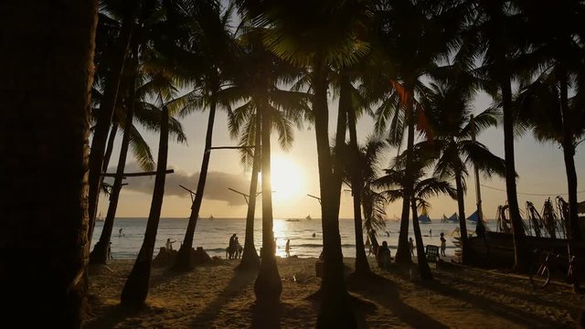 Beach with silhouettes of tourists among palm trees on the island of Boracay. Palm trees in the rays of sunset. Sailboats on the water. Philippine Tropics. Shooting in motion.