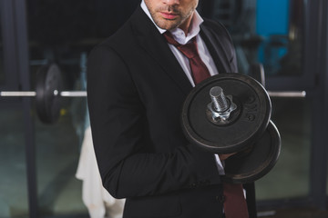 cropped view of sporty businessman in suit training with dumbbell in gym