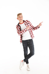 Full length portrait of happy educated teenage boy 17y wearing plaid shirt smiling and pointing fingers aside on copyspace, isolated over white background