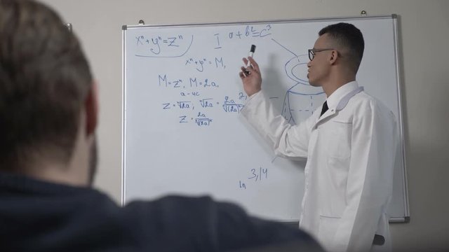 A young successful African scientist in a white coat and glasses, stands at the blackboard with formulas, presentation, lecture, training concept. 60 fps