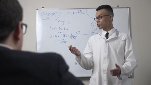 A young successful African scientist in a white coat and glasses, stands at the blackboard with formulas, presentation, lecture, training concept. 60 fps