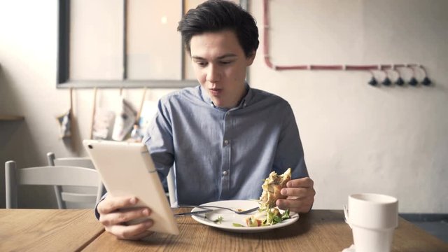Young teenager with tablet computer eating sandwich in cafe
