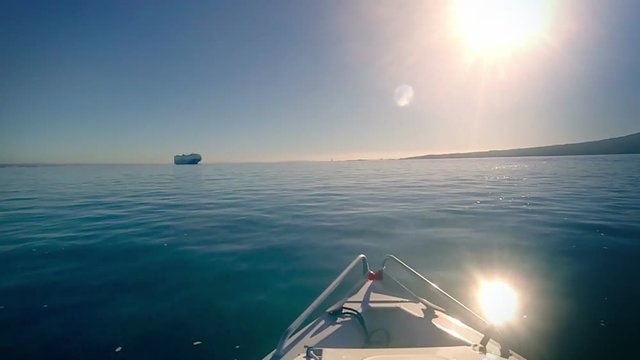 Motor boat onboard view, travelling through calm seas. The sun is reflected in the water. Location: Hauraki Gulf, New Zealand. Rangitoto Island at right. A cargo container ship is in the distance. 