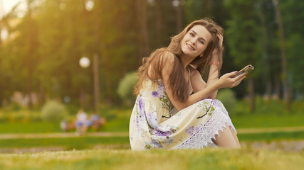 Young beautiful woman in summer dress with long hair sitting on grass in green park and talking on the phone, smiling. Summer. Recreation. Youth.