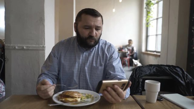 Young man watching movie on smartphone and eating meal sitting in cafe  
