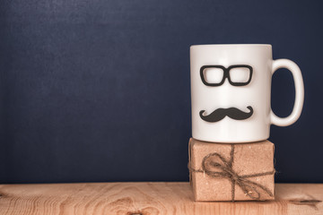 cup with mustache, glasses, gift box over black chalkboard, holiday concept of Fathers Day