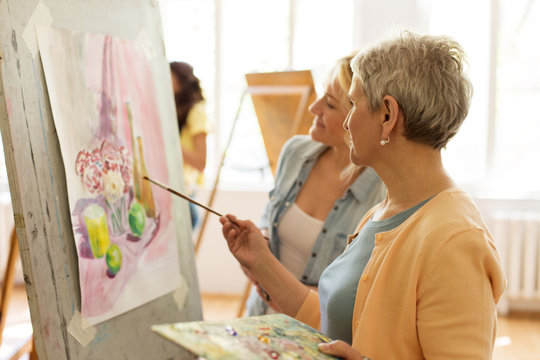 creativity, education and people concept - female artists or students with brushes and palettes painting on easel at art school studio